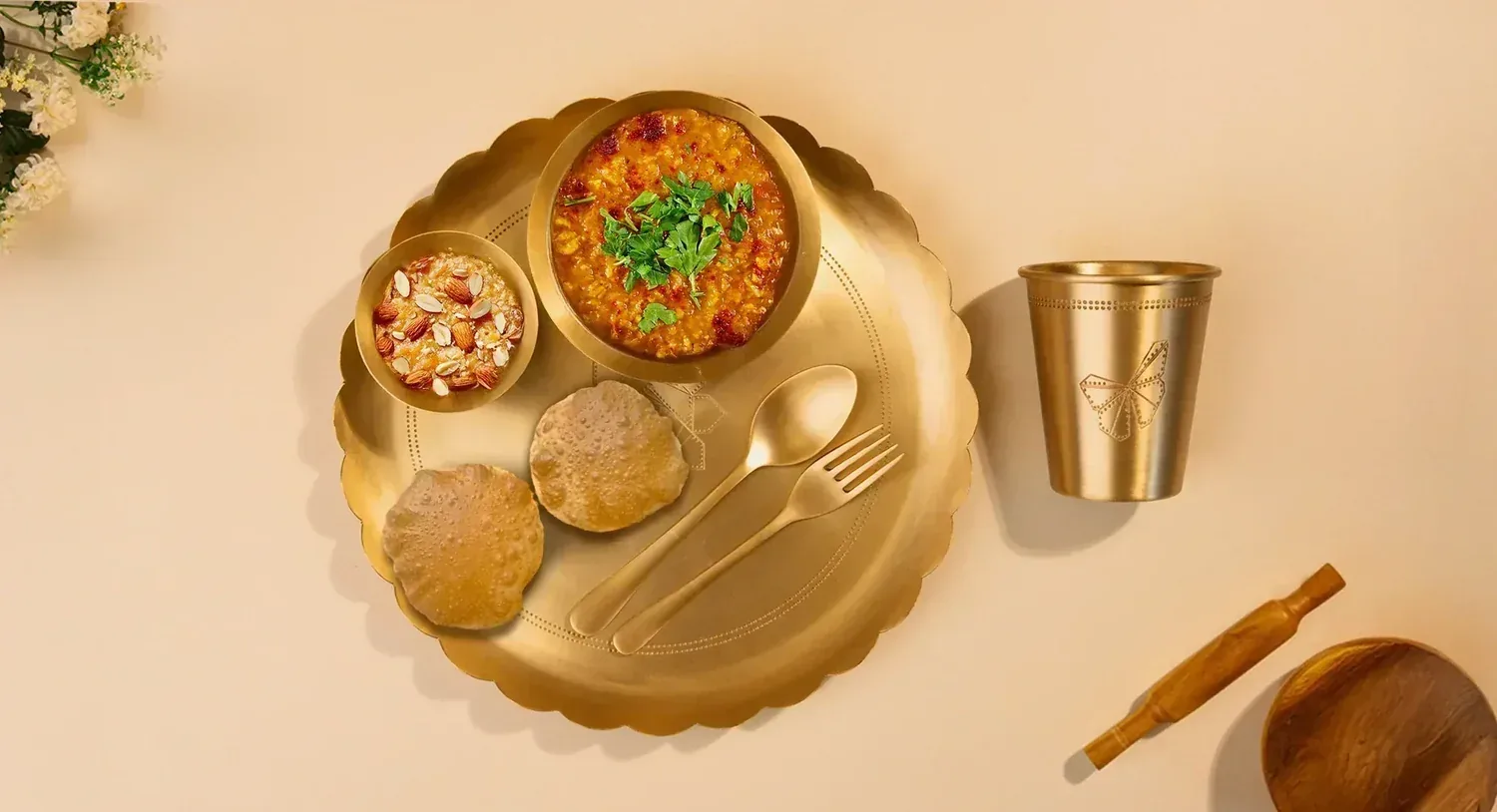 Brass thali with a bowl of dal, almond sweet, two puris, spoon and fork beside a brass tumbler for the Annaprashana first food ceremony
