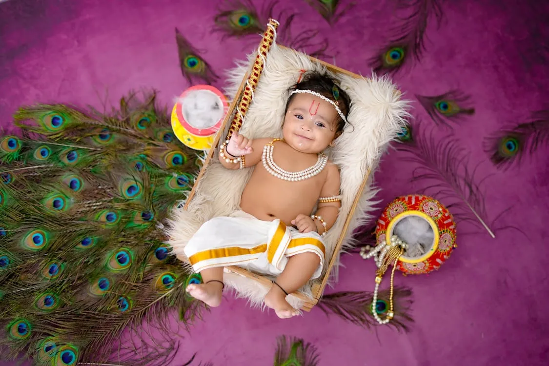 Smiling baby dressed in a white dhoti with pearl jewellery and a tilak, lying in a wooden crate on fur and surrounded by peacock feathers and decorated pots