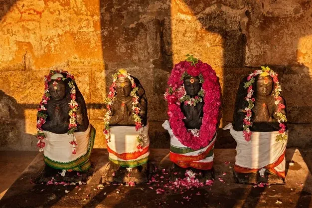 Three dark stone statues with folded hands, decorated with flower garlands and white and green cloth, in a sunlit temple