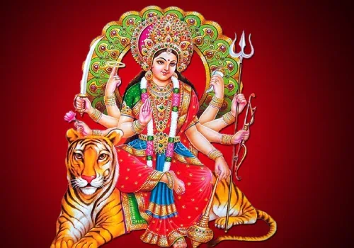 Goddess Durga with eight arms holding weapons and a trident, seated on a tiger against a deep red background