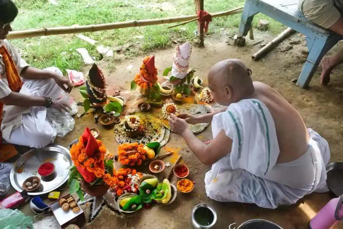 Man in a white dhoti arranging flower-decorated kalash, fruits and offerings on the ground during a Hindu puja