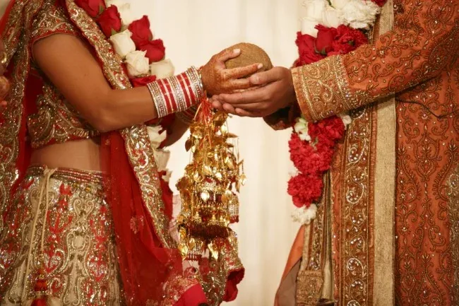 Bride and groom in red and gold wedding attire and flower garlands holding a coconut together during a Hindu wedding ritual
