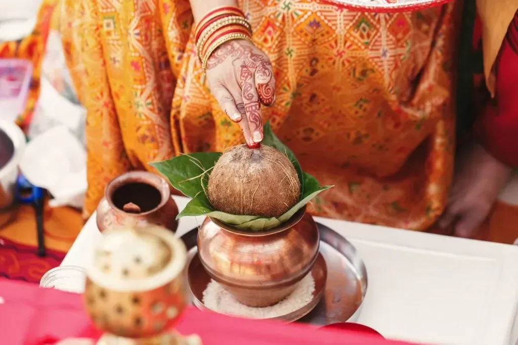 Hand decorated with henna applying red kumkum to a coconut on betel leaves resting on a copper kalash during a puja