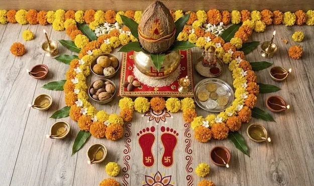 Puja setup with a coconut-topped kalash on a red cloth, surrounded by marigold garlands, oil lamps, nuts, coins and red Lakshmi footprints