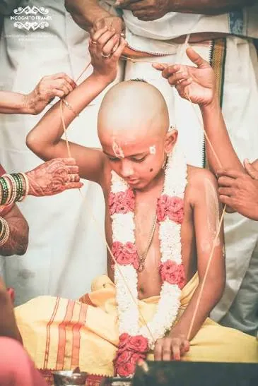 Boy with a shaved head and flower garland in a yellow dhoti as family hands hold a sacred thread around him during a Hindu ceremony